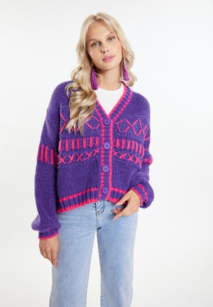 Lila gestrickte Strickjacke mit rosa Akzenten, einem V-Ausschnitt und fünf Knöpfen. Enthält geometrische Muster und weite Ärmel. Kombiniert mit hellen Jeans.
