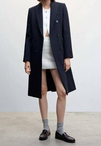 Cappotto di lana navy doppiopetto con rever a dischi, abbinato a un top corto in maglia bianca e una gonna testurizzata, calzini grigi e mocassini scuri.