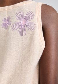Haut sans manches en maille crème avec des motifs floraux brodés violets près de l'épaule, porté par une personne à la peau foncée.