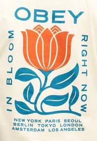 Έντονο πορτοκαλί λουλούδι με μπλε στέλεχος και φύλλα, περιβαλλόμενο από μπλε κείμενο: «ΥΠΑΚΟΥΣΕ», «ΣΕ ΑΝΘΙΣΗ», «ΑΥΤΗ ΤΗ ΣΤΙΓΜΗ» και ονόματα πόλεων παρακάτω.