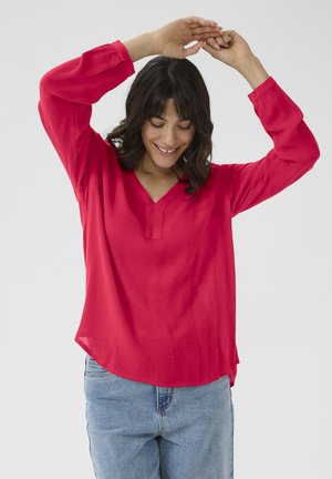 Blouse rouge à manches longues avec un col en V et des poignets boutonnés, dotée d'un tissu texturé. Associée à un jean bleu clair.