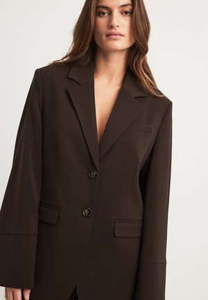 Bruine op maat gemaakte blazer met een diepe V-hals, twee voorvakken en een sluiting met twee knopen. Gemaakt van een soepele stof met brede mouwen.
