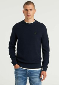 Marineblaues Strickpullover mit gerippter Textur, Rundhalsausschnitt und kleinem Logopatch auf der Brust, kombiniert mit hellblauen Jeans.