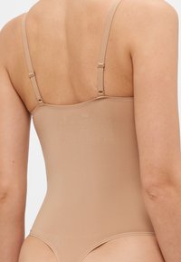 Body moldeador beige con tirantes ajustables y textura suave. Presenta un diseño sin costuras y un branding minimalista en la parte posterior.