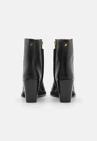 Botas de cuero negro hasta el tobillo con un acabado brillante, tacones altos y gruesos, y cremalleras doradas en la parte trasera. Punta redondeada y detalle de costura en los lados.