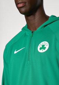 Nærbillede af en person iført en grøn Celtics half-zip hoodie med et hvidt Nike-logo på brystet.