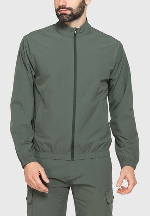 Uomo che indossa giacca verde oliva con zip e pantaloni cargo abbinati, in piedi davanti a uno sfondo chiaro e uniforme.