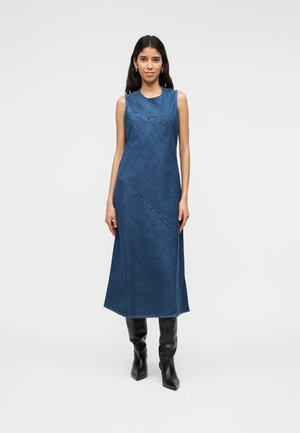 Kobieta stojąca w niebieskiej, bezrękawowej sukience midi z denimu i czarnych kozakach na obcasie do kolan na jednolitym tle.