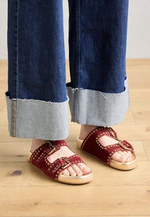 Pieds chaussés de sandales en daim rouge avec boucles et œillets dorés, associés à un jean à jambes larges retroussé aux chevilles, debout sur un sol en bois.