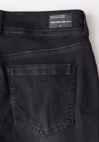 Černé džíny z denimu s texturovaným povrchem, s zadní kapsou a štítkem přišitým na pase obsahujícím pokyny pro úpravu velikosti a údržbu.