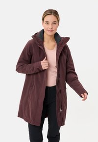 Vrouw die de rits van een lange bordeauxrode jas met donkere voering aanpast, gekleed in een lichtroze top en zwarte broek, staand op een witte achtergrond.