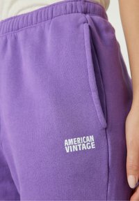 Violetinės sportinės kelnės, pagamintos iš minkštos medžiagos, su šoninėmis kišenėmis ir balta logo, kuri užrašyta "AMERICAN VINTAGE" priekyje.