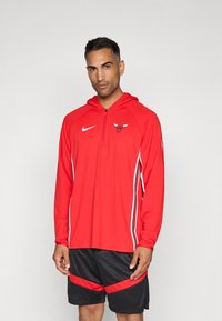 Rød hettegenser med glidelås krage, med Chicago Bulls-logo og hvite sidestriper, kombinert med svarte shorts med røde detaljer.
