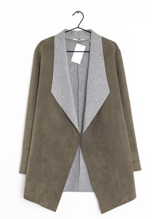 Veste en daim marron ouverte sur le devant avec de larges revers en tissu gris, manches longues et une étiquette blanche, suspendue sur un cintre noir.