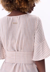 Gestreepte jurk van zacht materiaal, met lichtbeige en bruine strepen, uitlopende mouwen, een getailleerde taille, en een label met de tekst "blauwe schaduw."