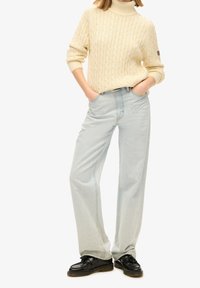Jeans dritti azzurro chiaro con un classico design a cinque tasche, abbinati a un maglione con collo a dolcevita in maglia a treccia color crema. Mocassini neri completano il look.