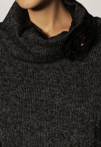 Svart, texturerad cowl-neck tröja med ett subtilt mönster. Har en tygblomma i svart med en orange mitten som accent.