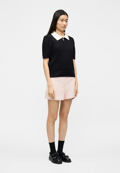 Femme debout portant un pull à manches courtes texturé noir avec un col blanc, un short rose clair, des chaussettes noires et des mocassins noirs.