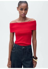 Rode off-shoulder top van rekbare stof, gecombineerd met donkerdenim high-waisted broek. Accessoires omvatten gestapelde armbanden.