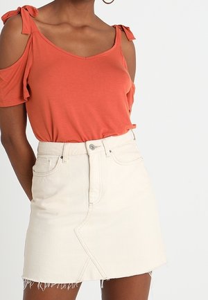 Roest-oranje cold-shoulder top met strikaccenten, gecombineerd met een witte denim minirok met een rafelige zoom en diagonale naaddetails.