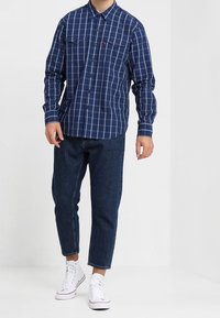 Chemise à carreaux bleu marine, à boutons, manches longues, avec deux poches poitrine ; associée à un jean en denim noir légèrement court et des baskets blanches.