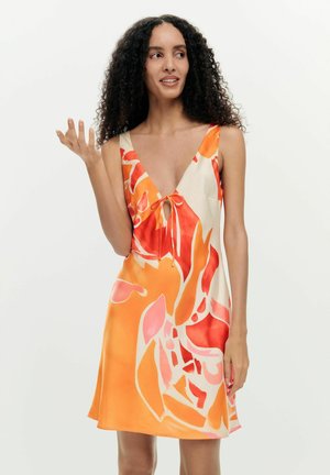 Woman with curly hair wearing sleeveless orange and red abstract patterned dress, gesturing with one hand, standing against white background.