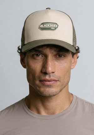 Man with short curly hair wears beige and olive cap with "BLACKSKIES PRO" logo and a matching beige crew neck shirt, neutral background.