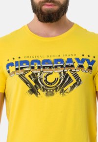 Yellow cotton t-shirt featuring a graphic print with blue and black text "CIPO & BAXX" and mechanical design elements on the front.