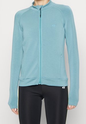 Kobieta ubrana w jasnoniebieską kurtkę zapinaną na zamek z długimi rękawami oraz czarne legginsy z małym białym logo w pobliżu biodra.