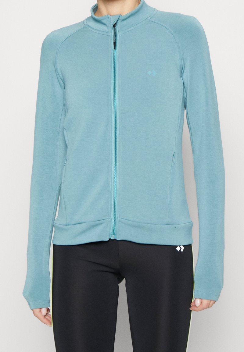 Femme portant une veste zippée bleu clair à manches longues et un legging noir avec un petit logo blanc près de la hanche.