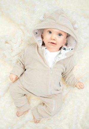 Jasnobeżowy kombinezon z polaru z suwakiem, z kapturem ozdobionym uszami, podszyty tkaniną w wzory. Miękka tekstura, długie rękawy i widoczne mankiety.