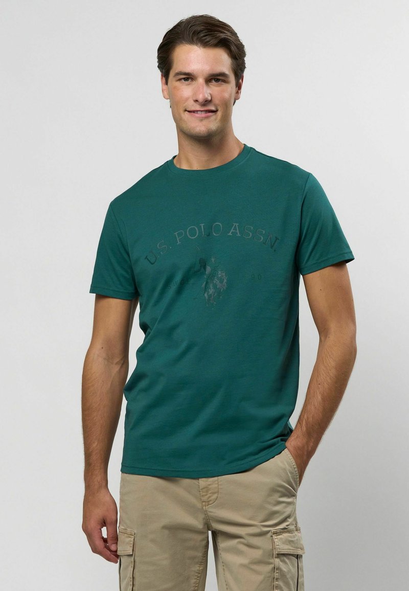 Tyrkysové bavlnené tričko s crew výstrihom a krátkymi rukávmi, s potlačeným logom v tmavšej farbe na prednej strane.