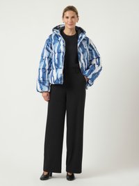 Moteris, vilkinti mėlyną ir baltą raštuotą pūkinę striukę, juodą ilgomis rankovėmis marškinėlį, plačius juodus kelnes ir juodus kulnus, stovi su viena ranka kišenėje.