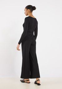 Black long-sleeve jumpsuit with a fitted bodice and wide-leg trousers, featuring a low back, made from smooth fabric, paired with black sandals.