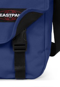 Modrá textilní taška přes rameno s černým klopovým víkem, sponou na uzávěr a s nášivkou loga Eastpak. Materiál má texturovaný povrch.