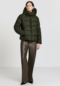 Giacca a vento imbottita verde oliva con collo alto, cerniera frontale e pannelli cuciti, abbinata a pantaloni larghi lucidi verde oliva e scarpe nere.