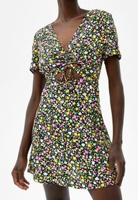 Krótka sukienka w kwiaty z czarnym tłem, zdobiona kolorowymi różowymi, żółtymi i zielonymi kwiatami. Ma dekolt w kształcie litery V, krótkie rękawy oraz wycięcia.