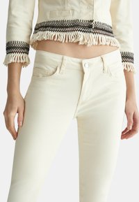 Chupa de denim color crema con detalles de flecos y acentos bordados en negro en los puños, combinada con unos jeans ceñidos color crema a juego.