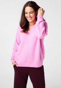 Roze V-hals trui met ribbelstructuur en verlaagde schouders, gecombineerd met donker pantalon. De stof lijkt zacht en lichtgewicht.