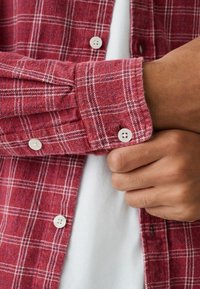 Rotes kariertes Flanellhemd mit weißen Knöpfen, einem strukturierten Gewebe und sichtbaren Manschettendetails, über einem weißen T-Shirt getragen.