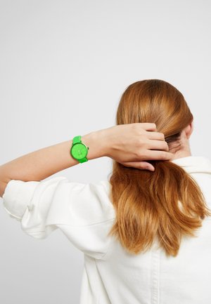 Personne aux longs cheveux rouges portant une chemise blanche, retenant ses cheveux en arrière, portant une montre-bracelet vert vif sur le bras gauche, sur un fond uni.