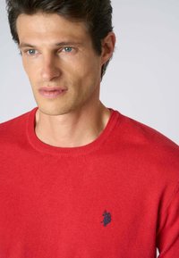 Maglione rosso a maglia con scollatura rotonda, con un logo ricamato blu sul lato sinistro del petto. Texture morbida, design minimalista.