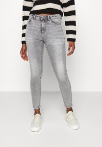 Jeans skinny gris avec un look usé, présentant une taille mi-haute, des passants de ceinture et un design classique à cinq poches. Associés à des baskets blanches.