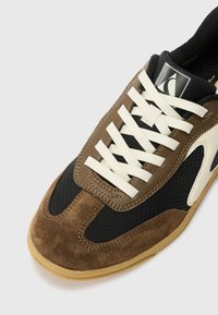 Brown and black sneaker with a textured design, suede and mesh materials, white laces, and a rubber sole. Features logo on the tongue.