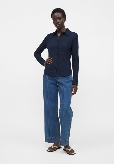 Femme portant une chemise bleu marine à manches longues boutonnée, un jean bleu à jambes larges avec des détails de couture à l’avant, et des tongs beiges, debout avec une main sur la hanche.