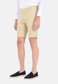 Pantaloni corti beige, lunghi fino al ginocchio, realizzati in un tessuto testurizzato, con fronte piatto, tasche laterali e risvolti. Abbinati a scarpe navy senza lacci.