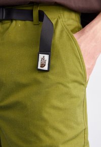 Olivgrön byxor tillverkade av slitstarkt tyg, med en svart bältesögla och en märkesetikett, slät textur och klassisk passform.