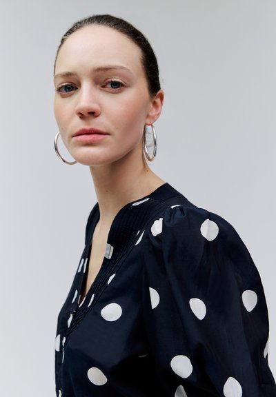 Világos bőrű, kék szemű nő nagy ezüst karika fülbevalóval és fekete, fehér pöttyös blúzzal, a kamerába nézve.