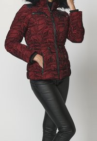 Kobieta w czerwonej kurtce puchowej w wzory, z czarnym futrzanym kapturem i czarnymi skórzanymi spodniami, pozująca z jedną ręką w kieszeni, a drugą blisko twarzy.