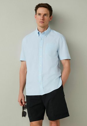 Hellblaues Kurzarm-Hemd mit Knopfleiste und Brusttasche, hergestellt aus einem glatten Stoff. Kombiniert mit schwarzen Shorts, Sonnenbrille in der Hand.